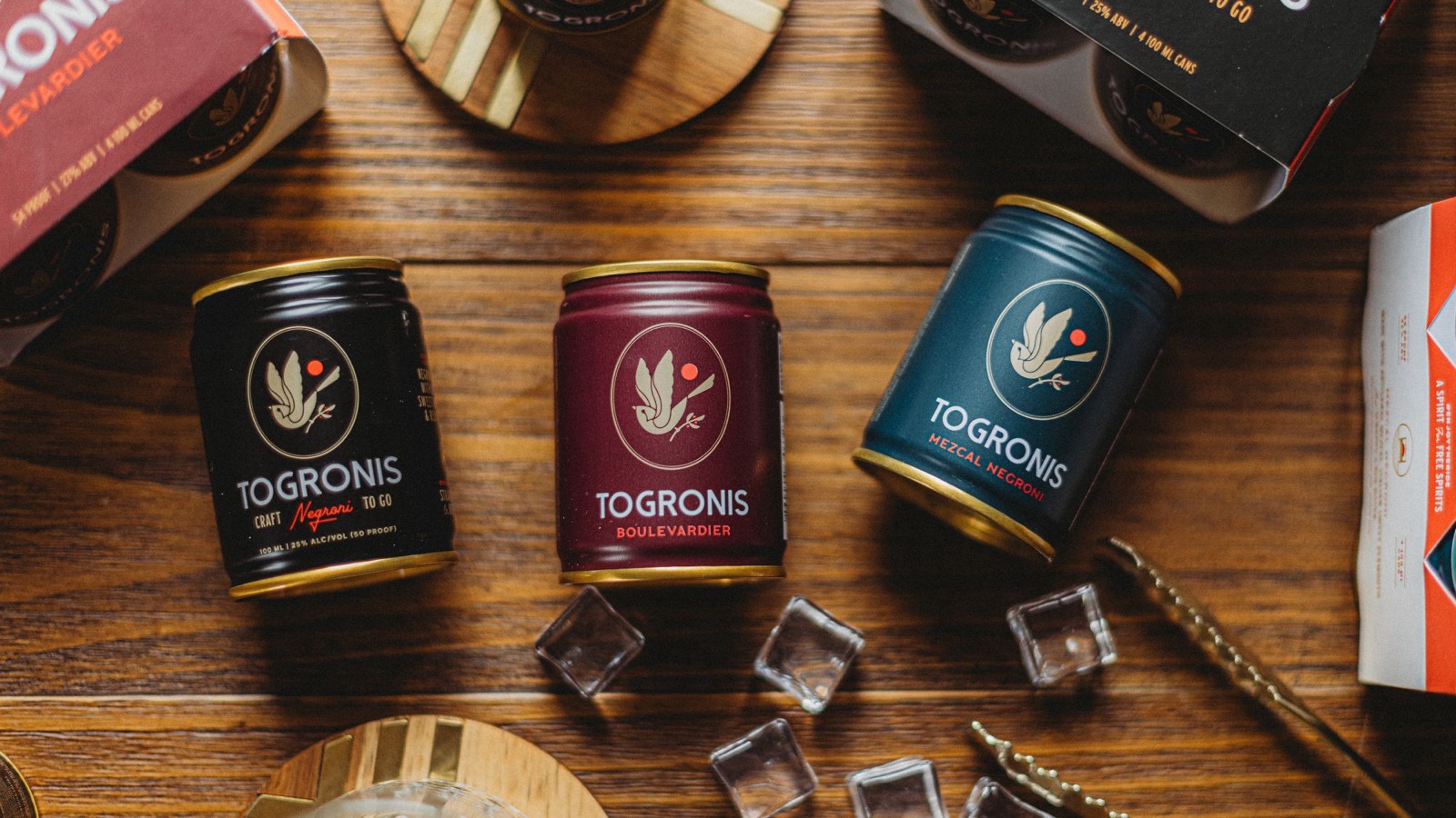 Togronis Negroni, Boulevardier and Mezcal Negroni cans on a wooden bar with ice and their four-pack boxes